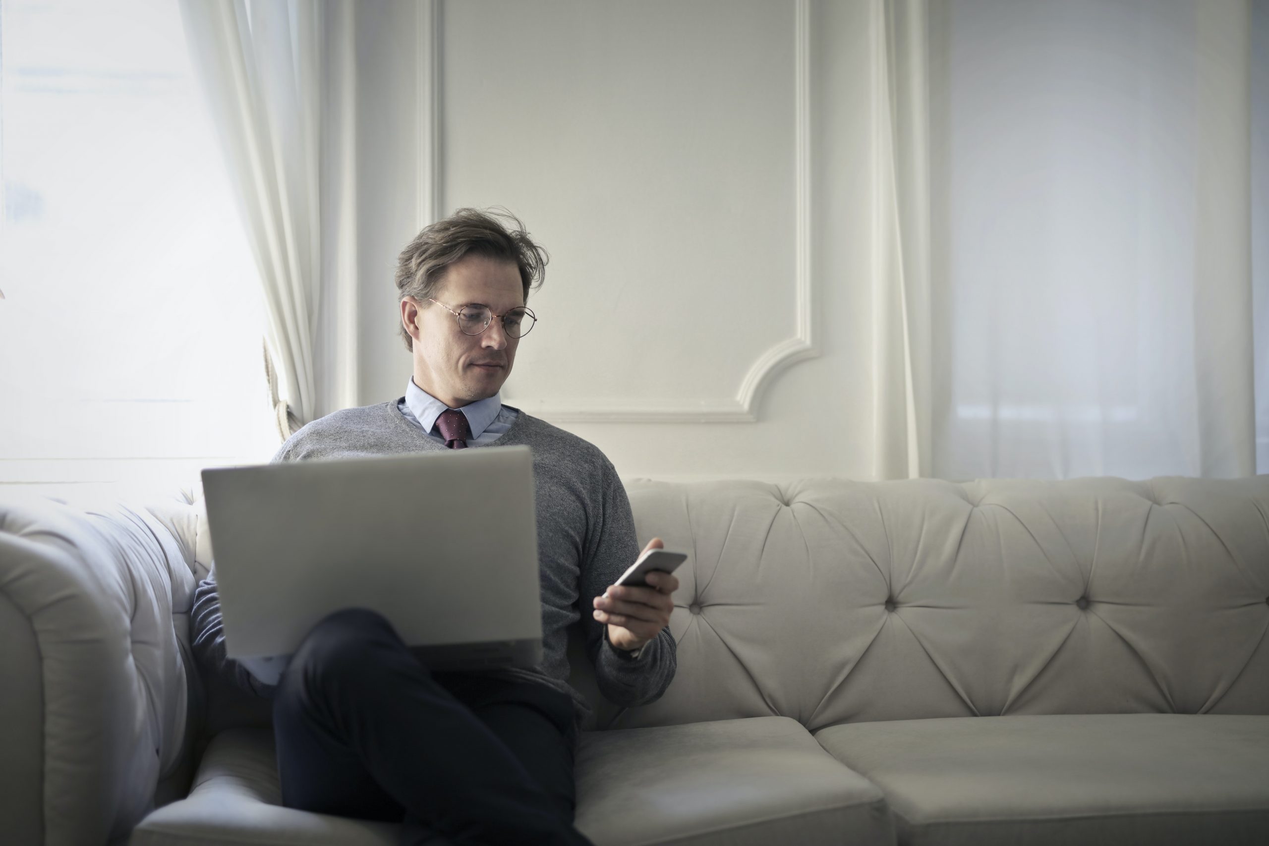 Nachdenklicher Geschäftsmann im eleganten Anzug sitzt auf einem Sofa, betrachtet ein Smartphone und arbeitet an einem Laptop, möglicherweise um Versicherungsoptionen online zu vergleichen oder eine Versicherungspolice zu verwalten, in einem hellen, stilvollen Raum.