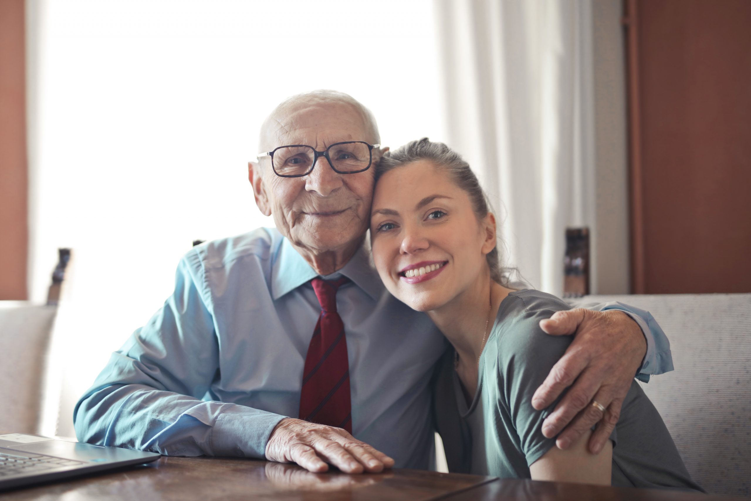 Ein älterer Mann mit Brille und Krawatte sitzt neben einer jüngeren Frau, die ihn umarmt, während sie beide lächeln und vor einem Laptop sitzen, was vertrauensvolle Finanzberatung zwischen Generationen veranschaulicht.