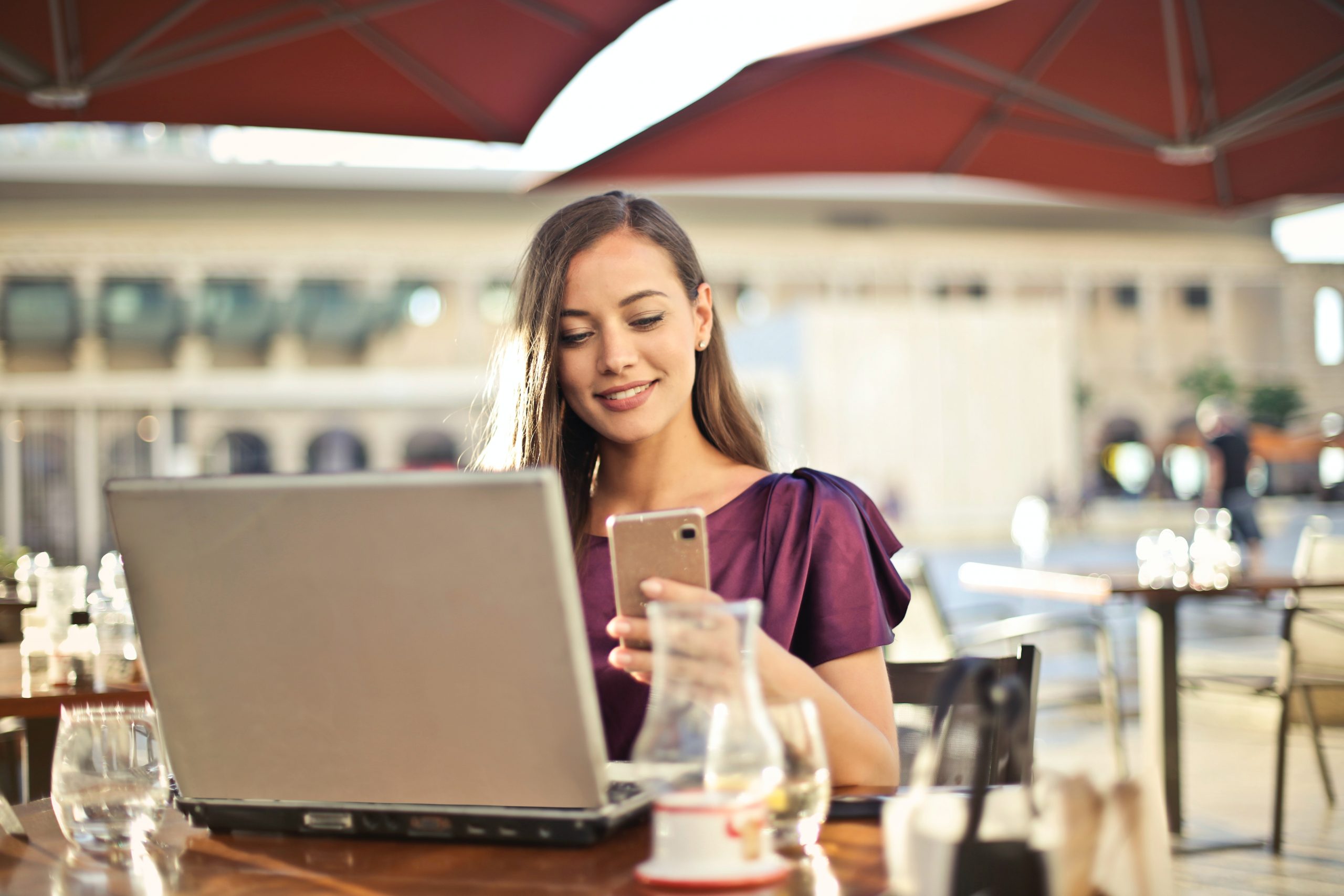 Eine konzentrierte junge Frau in einem pflaumenfarbenen Oberteil, die an einem Außentisch eines Cafés auf ihr Smartphone schaut und gleichzeitig an ihrem Laptop arbeitet, möglicherweise um Online-Kredite zu recherchieren oder ihre Finanzen zu verwalten.