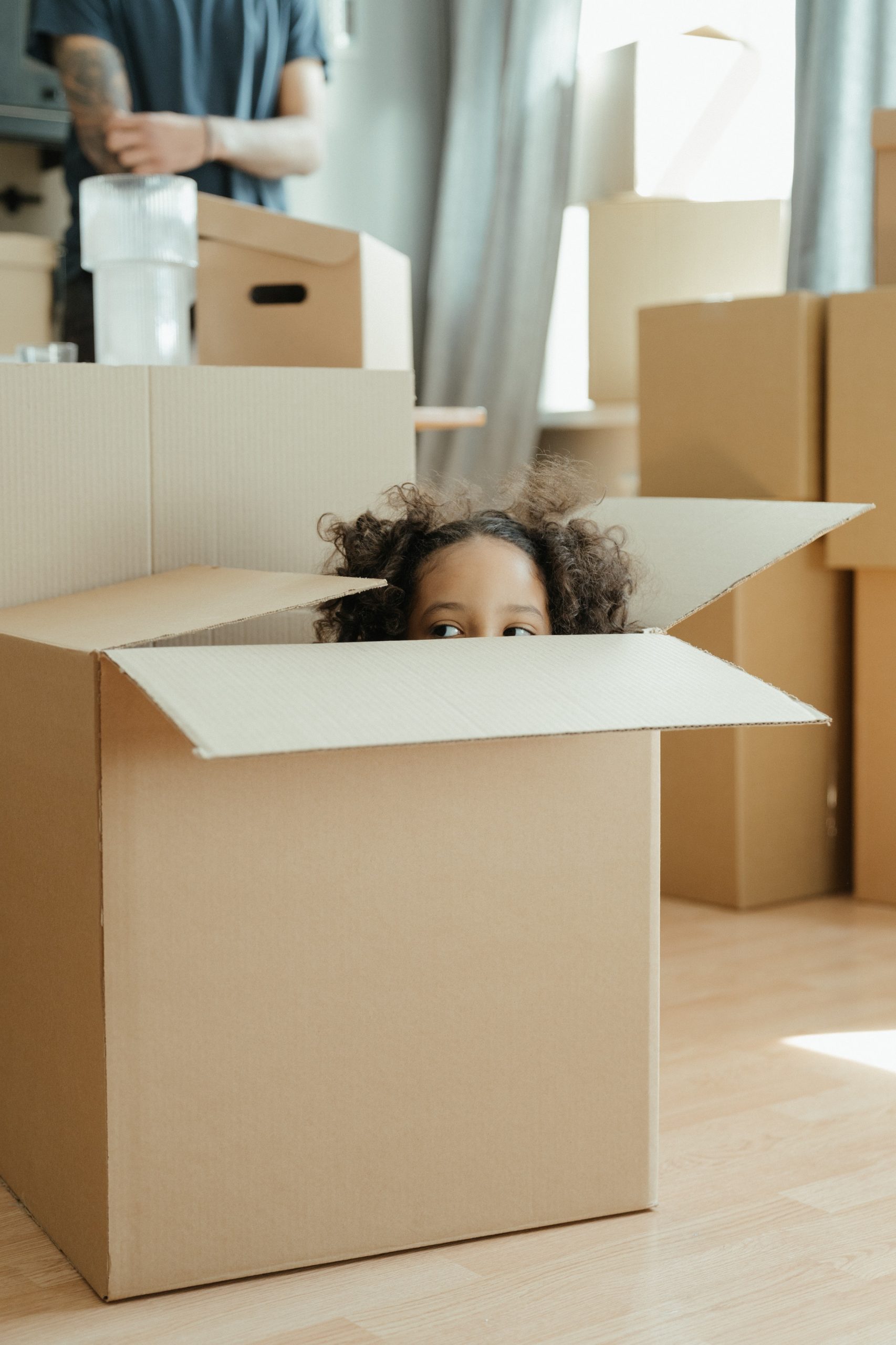 Ein Kind, das aus einer Umzugskiste schaut, während im Hintergrund Erwachsene mit weiteren Kisten beschäftigt sind, veranschaulicht die Wichtigkeit einer Hausratsversicherung beim Schutz von Besitztümern während eines Wohnungswechsels.