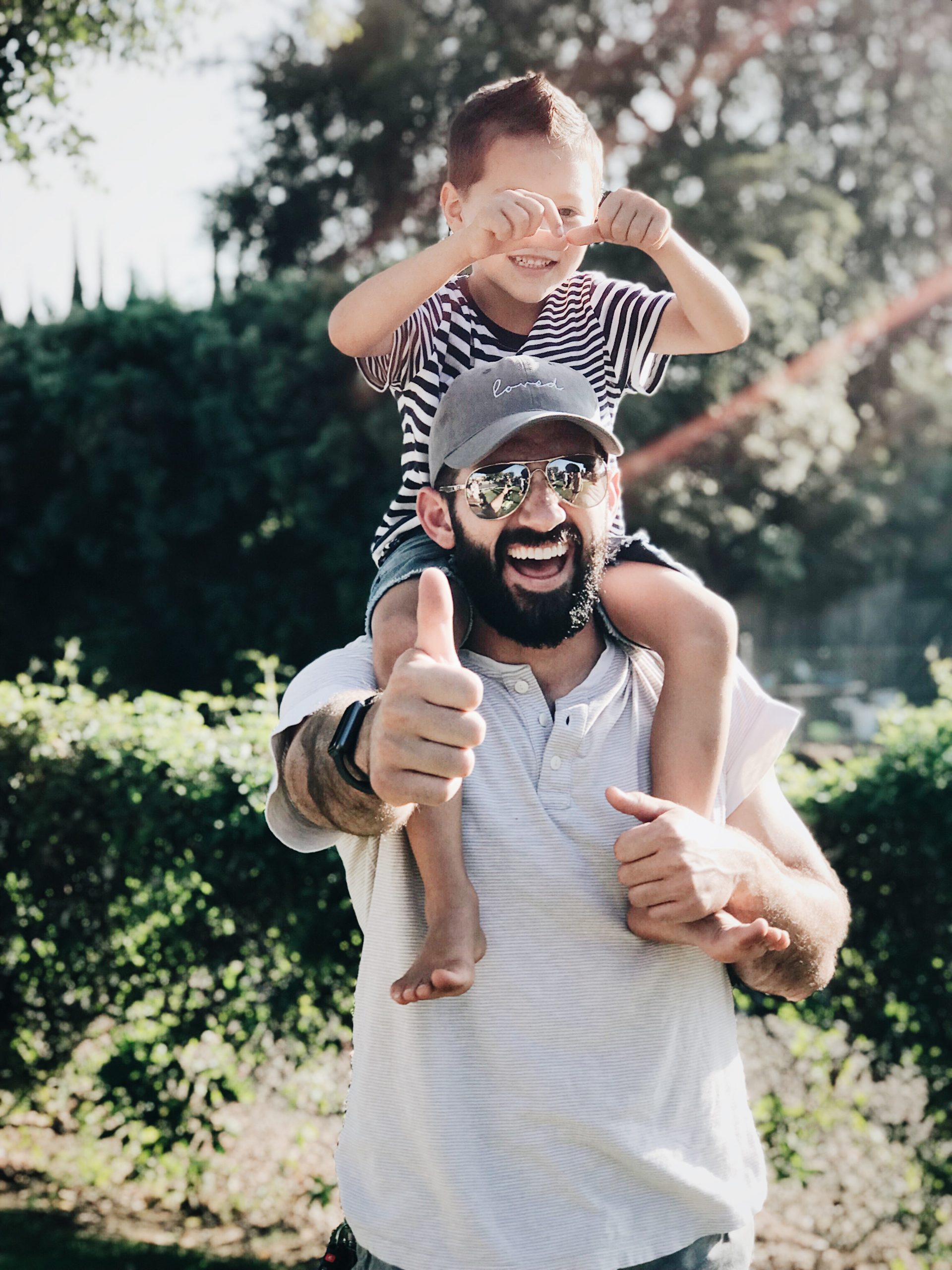 Fröhlicher Vater trägt sein Kind auf den Schultern im Freien und beide zeigen Daumen hoch, was die Freude und Sicherheit symbolisiert, die Familien durch zuverlässige Versicherungen genießen können.