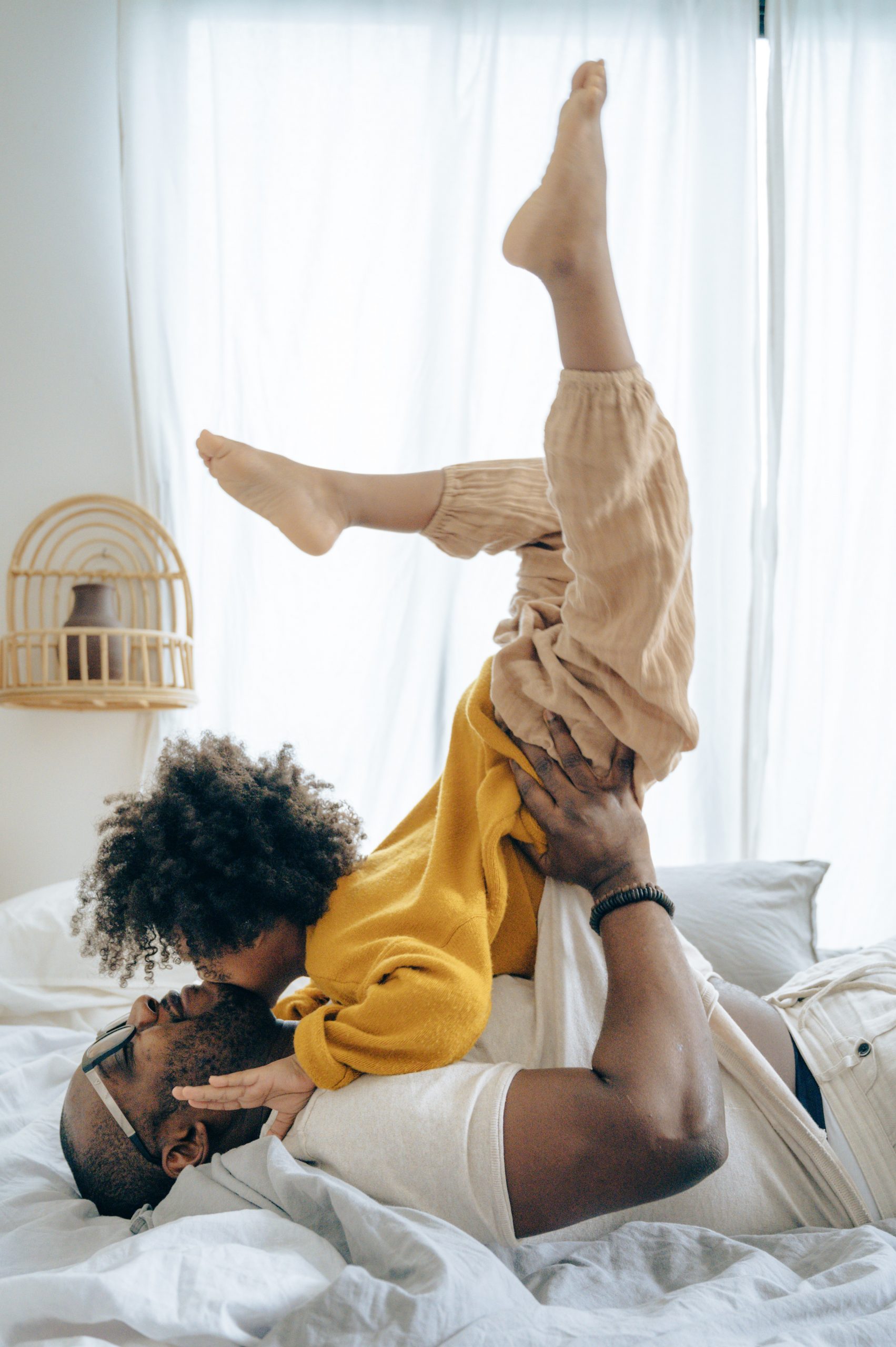 Ein fröhlicher afroamerikanischer Vater spielt zu Hause mit seinem Kind auf dem Bett, was die Bedeutung einer soliden Altersvorsorgeplanung für die Zukunft und das Wohlergehen der Familie unterstreicht, in einem hellen, gemütlichen Schlafzimmer.